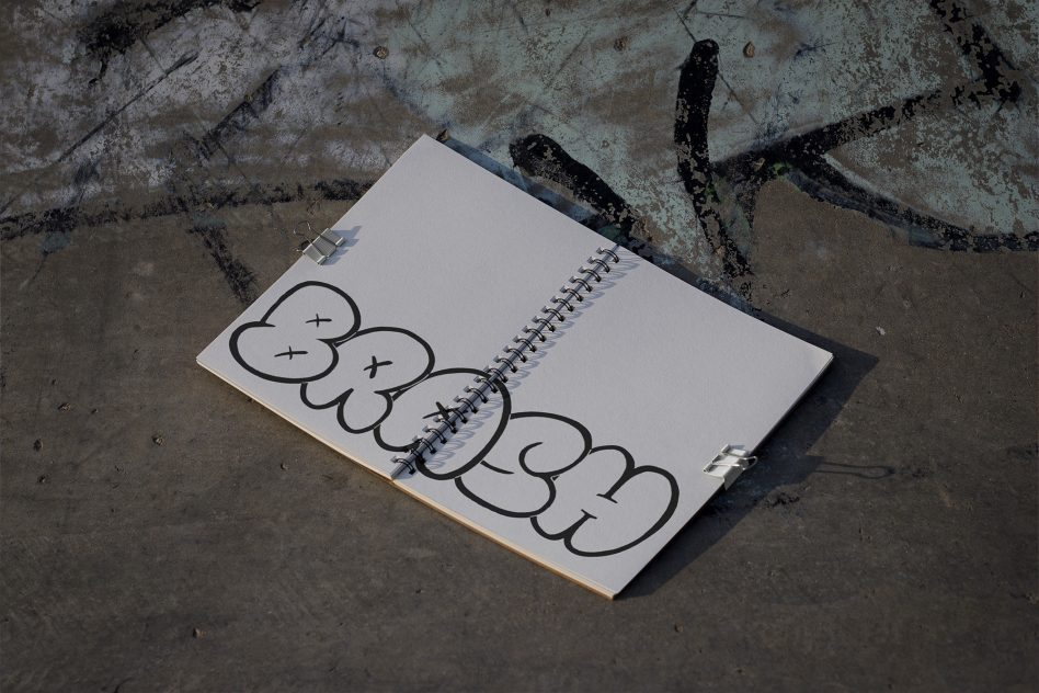 Spiral notebook mockup with artistic sketch on textured street background, showcasing graffiti style font design in a creative urban setting.